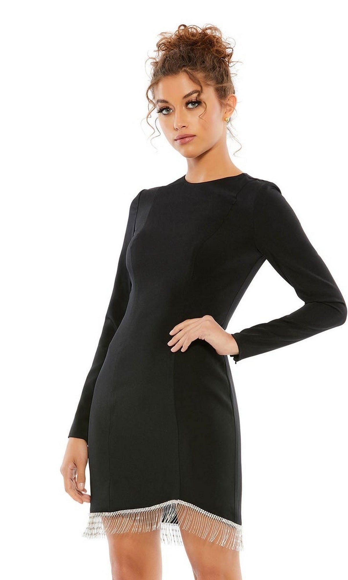 A woman wears the Short Homecoming Dress 26679 by Mac Duggal—a fitted black party dress with long sleeves and a beaded fringe hem—standing confidently with one hand on her hip against a plain background.